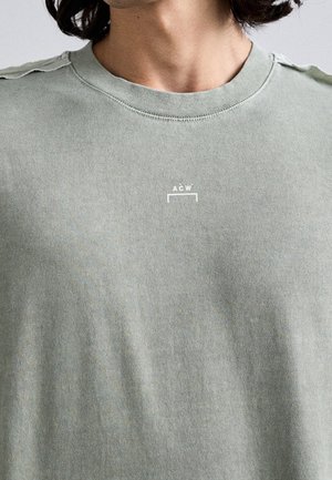 Personne portant un t-shirt vert clair à col rond avec un petit logo blanc "A C W" centré sur la poitrine.