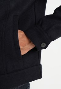 Granatowa kurtka z mieszanki wełny z teksturowanym materiałem, wyraźną kieszenią boczną oraz kontrastowym, czarnym detalem guzikowym przy mankietach.