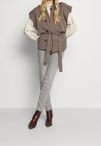 Chaleco sin mangas color topo con gran cuello y bolsillos frontales, cintura ceñida con cinturón, combinado con jeans skinny gris claros y botas de tobillo puntiagudas marrones.