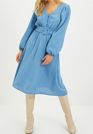 Kobieta w niebieskiej sukience midi z długimi bufiastymi rękawami, zapięciem na guziki z przodu, szerokim paskiem i beżowymi kozakami do kolan, stojąca na białym tle.