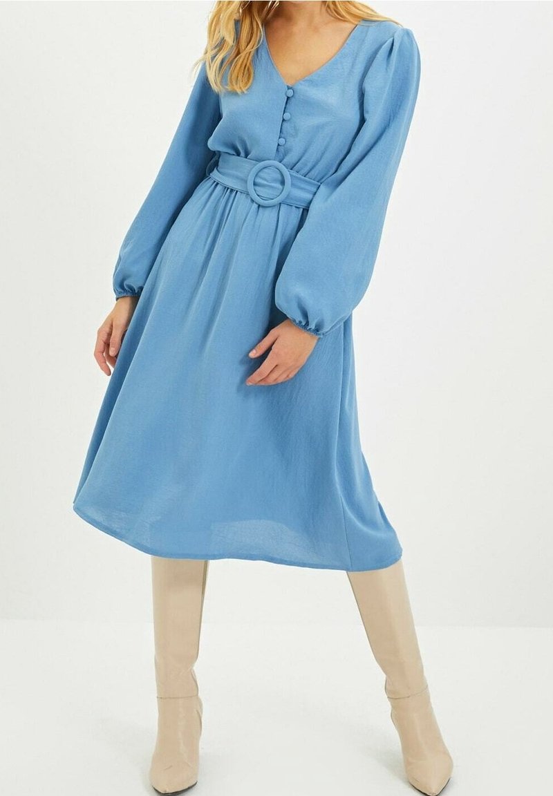Femme portant une robe midi bleue avec de longues manches bouffantes, devant boutonné, ceinture large et bottes beige hauteur genoux, debout sur fond blanc.