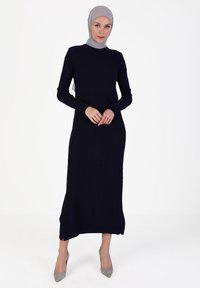 Long navy ribbed dress with a high neckline and long sleeves, featuring side slits at the hem. Accompanied by a light gray hijab and heels.