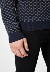 Manlig hand vilande bredvid benet iförd en marinblå tröja med vita snöflingemönster och svarta byxor.