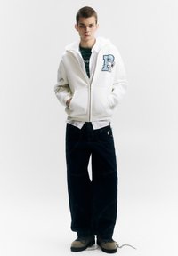 Witte capuchontrui met ritssluiting, voorzakken en een blauw letterpatch; donkere wijde jeans met zichtbare stiksels; grijze schoenen met blauwe accenten.