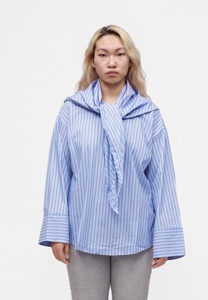 Giovane con capelli biondi lunghi che indossa una camicia blu a righe oversize e un maglione appoggiato sulle spalle, in piedi davanti a uno sfondo bianco.
