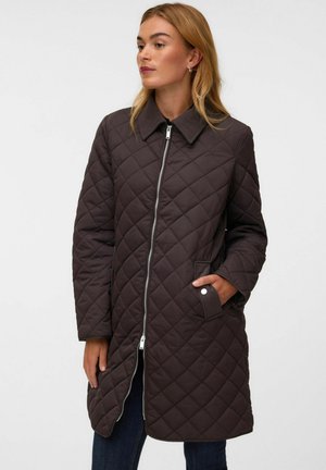 Femme portant un long manteau matelassé marron foncé avec une fermeture éclair à l'avant et un col, une main dans une poche à bouton-pression.