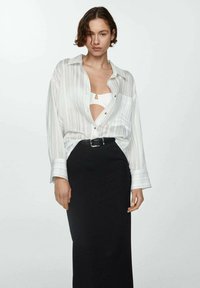 Vrouw met kort bruin haar, gekleed in een witte gestreepte blouse die open is over een witte top, in een hooggetailleerde zwarte rok met een riem.