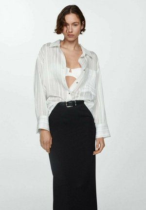 Vrouw met kort bruin haar, gekleed in een witte gestreepte blouse die open is over een witte top, in een hooggetailleerde zwarte rok met een riem.