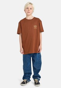 Braunes Baumwoll-T-Shirt mit rundem Halsausschnitt und kurzen Ärmeln, versehen mit einem grafischen Design in Gelb auf der Brust. Getragen mit blauen Jeans.