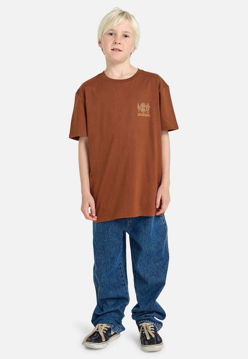 Braunes Baumwoll-T-Shirt mit rundem Halsausschnitt und kurzen Ärmeln, versehen mit einem grafischen Design in Gelb auf der Brust. Getragen mit blauen Jeans.