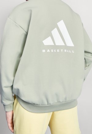 Persoon die een lichtgroene sweatshirt draagt met een groot wit Adidas-logo en het woord "BASKETBAL" op de achterkant, gecombineerd met bleekgele broek.