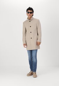 Abrigo de mezcla de lana beige con cuello alto, botones frontales y bolsillos laterales, combinado con vaqueros azules y zapatos marrones. Modelo lleva gafas de sol.