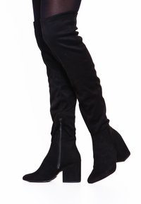 Černé kozačky nad kolena vyrobené z měkké semišové kůže s špičatou špičkou, boční zipovou uzávěrkou a robustním podpatkem pro dodatečnou stabilitu.