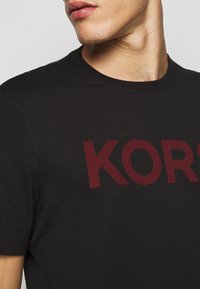Černé tričko s kulatým výstřihem s velkým burgundským grafickým potiskem "KORS" na přední straně. Vyrobeno z měkké látky s hladkým povrchem.