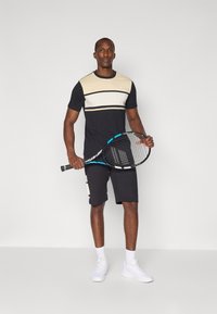Conjunto deportivo de hombre que presenta una camiseta a rayas en negro y beis con bloques de color y pantalones cortos negros. Sosteniendo una raqueta de tenis. Zapatillas blancas.