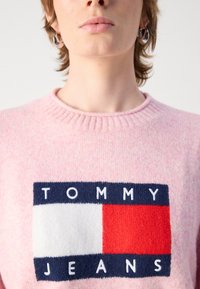 Rožinė megzta suknelė su dideliu „TOMMY JEANS“ logotipu tamsiai mėlynos ir raudonos spalvos, su išraižyta apykakle ir minkšta tekstūra.