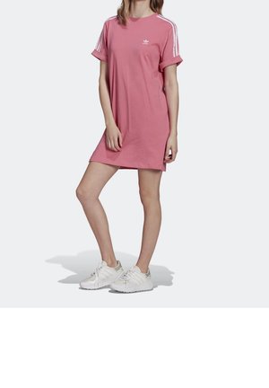 Vrouw draagt een roze Adidas T-shirtjurk met witte strepen op de mouwen en witte sneakers, staand tegen een effen lichte achtergrond.