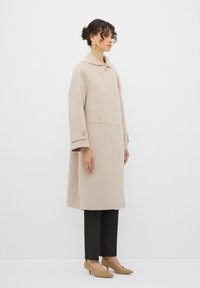 Cappotto di lana beige con colletto, grande bottone frontale e tasche laterali. Abbinato a pantaloni a righe e tacchi beige.