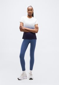 Γυναίκα φορώντας κοντομάνικη μπλούζα με διαβάθμιση από λευκό σε μαύρο, μπλε κολάν και λευκά αθλητικά παπούτσια, στέκεται με σταυρωμένα τα χέρια μπροστά σε λευκό φόντο.