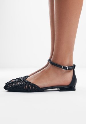 Scarpe nere piatte con materiale intrecciato sulla punta, un retro in pelle liscia e una sottile cinghia alla caviglia con una fibbia in metallo.