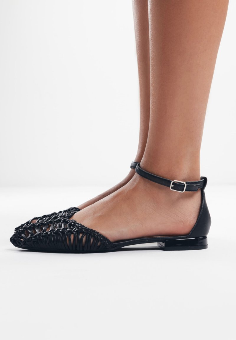 Zapatos planos negros con material trenzado en la punta, parte trasera de cuero suave y una tira delgada en el tobillo con una hebilla de metal.