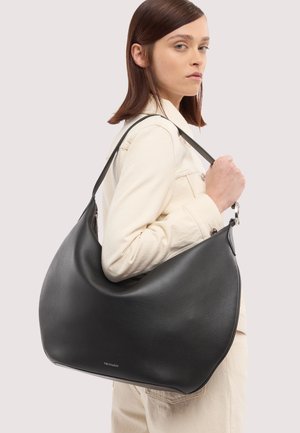 Woman wearing light beige jacket holding large black leather shoulder bag with curved shape and silver hardware.