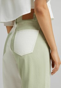 Pantalones bicolor en verde claro y blanco con un acabado texturizado, que presentan bolsillos de parche y una etiqueta de marca marrón en la cinturilla.