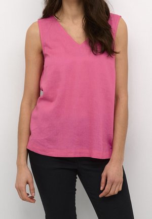 Nainen pukeutunut hihattomaan vaaleanpunaiseen V-kaulaiseen toppiin ja mustiin housuihin, seisoo yksinkertaisen vaalean taustan edessä.