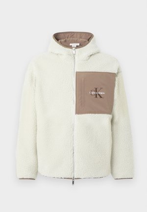 Fleece zip-jakke i off-white med en brun lomme. Har hette, teksturert stoff og "Calvin Klein"-logo på lommen.