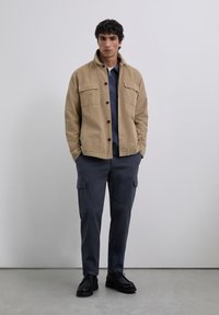 Joven vestido con una chaqueta beige abotonada, una camisa de cremallera azul marino, pantalones cargo oscuros y zapatos negros con cordones, de pie en el interior.