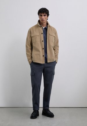 Junger Mann in einer beige geknöpften Jacke, einem marineblauen Reißverschlusshemd, dunklen Cargo-Hosen und schwarzen Schnürschuhen, der drinnen steht.