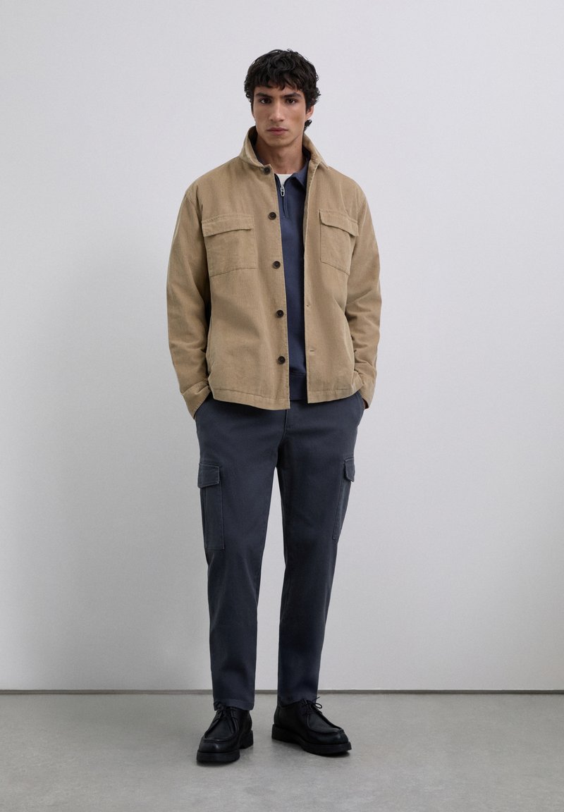 Joven vestido con una chaqueta beige abotonada, una camisa de cremallera azul marino, pantalones cargo oscuros y zapatos negros con cordones, de pie en el interior.