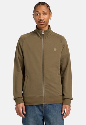 Mann mit geflochtenem Haar trägt olivgrüne Jacke mit Reißverschluss und blaue Jeans und steht vor einem schlichten hellgrauen Hintergrund.