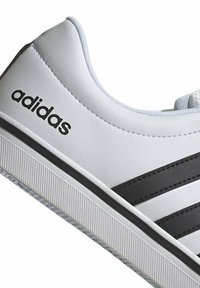 Zapatilla blanca con parte superior sintética texturizada, rayas negras y logotipo de "adidas". Cuenta con una suela de goma blanca con un patrón de diamantes.