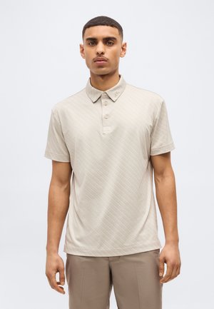 Tânăr cu păr scurt, purtând un tricou polo bej cu mâneci scurte și pantaloni maro deschis, stând pe un fundal alb.