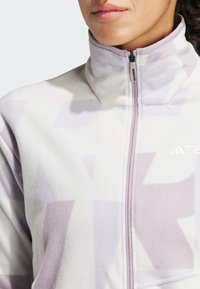 Chaqueta de forro polar en lavanda clara con patrón geométrico abstracto, cuello alto y cierre de cremallera. Textura suave con sutiles variaciones de color.