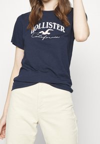 Námořnická modrá bavlněná tričko se znakem "Hollister California" v bílé a růžové barvě, kulatý výstřih a krátké rukávy, v kombinaci s krémovými sukněmi.