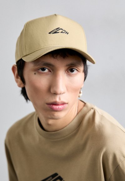 Casquette beige avec une visière incurvée, arborant un logo noir à l'avant. Le modèle porte un t-shirt beige assorti, sur un fond clair.