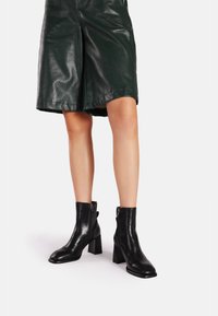 Stivaletti in pelle nera alla caviglia con punta quadrata, tacco a blocco e zip laterale, abbinati a pantaloni corti in pelle verde. Texture liscia e design elegante.