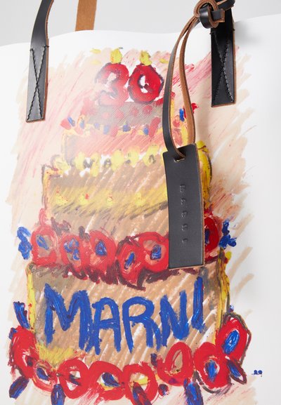 Sac fourre-tout avec une illustration colorée d'un gâteau d'anniversaire, avec des accents rouges et bleus, des textures brunes et des poignées en cuir avec une étiquette noire.
