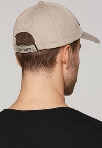 Cappellino beige da baseball con visiera curva. La fibbia regolabile sul retro presenta il logo "CAMP DAVID". La texture del tessuto appare liscia con cuciture visibili.