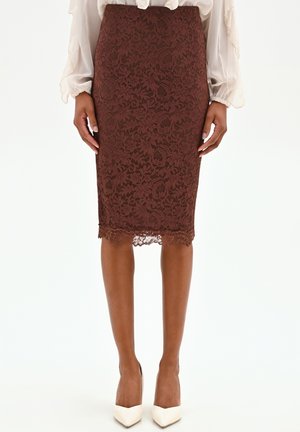 Gonna a matita in pizzo floreale marrone indossata con una camicetta trasparente a maniche lunghe e décolleté bianche con punta appuntita, vista dalla vita ai piedi.