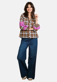 Camicia a quadri marrone con polsini ricamati rosa, chiusura a bottoni, maniche lunghe, abbinata a jeans in denim blu a gamba ampia e scarpe chiare.