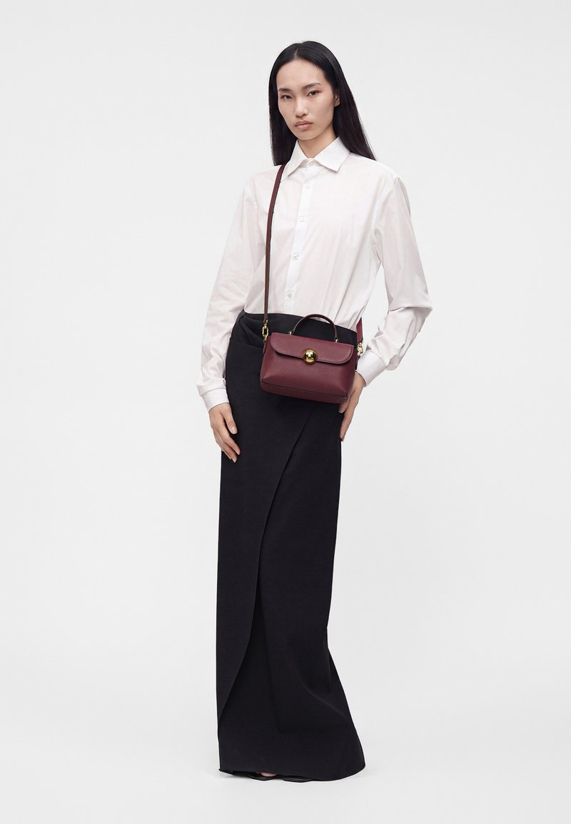 Femme aux longs cheveux noirs, portant une chemise blanche boutonnée, une jupe noire longue jusqu'au sol, et portant un petit sac à main rouge foncé avec une fermeture dorée.