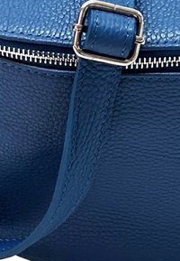 Blauwe leren tas met textuur, een zilveren rits en gouden hardware. Voorzien van een platte schouderband en een gladde oppervlak, wat een minimalistisch ontwerp benadrukt.