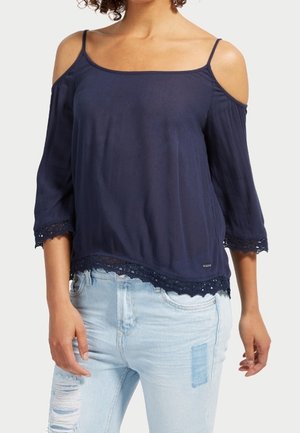 Kobieta ubrana w granatową bluzkę typu cold-shoulder z koronkowym wykończeniem oraz jasnoniebieskie podarte dżinsy, stojąca na tle jednolitego jasnego tła.
