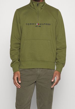 Man draagt een olijfgroene trui met een kwartrits en voorzak, met het logo "Tommy Hilfiger EST. 1985", gecombineerd met olijfgroene broek.