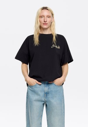 T-shirt nera oversize in cotone con maniche corte, che presenta un logo ricamato in oro sul lato sinistro del petto. Abbinata a jeans in denim blu chiaro.