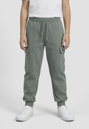 Bambino che indossa pantaloni jogger cargo grigi con polsini elastici e scarpe da ginnastica bianche, in piedi davanti a uno sfondo semplice.