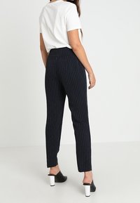 Pantalon noir à rayures avec une taille haute et des jambes fuselées, fabriqué en tissu lisse, associé à un t-shirt blanc et des chaussures à talons blocs noires.